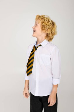 Okul üniformalı sarışın çocuk parlak gülümsüyor, dalgalı saçları ve çizgili kravatı var..