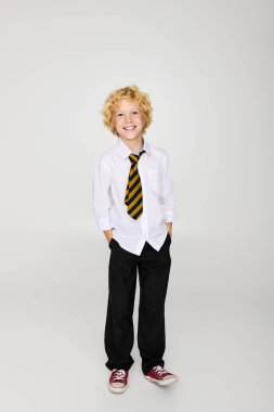 Dalgalı saçlı sarışın çocuk bir stüdyoda gülümserken okul üniformasıyla güvenle ayakta duruyor..