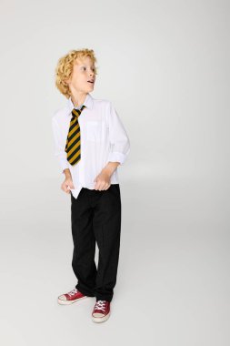 Okul üniformalı ve çizgili kravatlı dalgalı sarı saçlı neşeli bir çocuk stüdyoda duruyor..