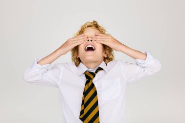 Okul üniformalı sarışın bir çocuk stüdyoda neşeyle gülerken gözlerini kapatıyor..