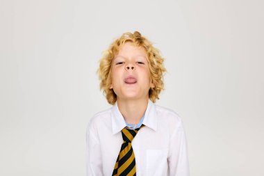 Beyaz gömlekli ve çizgili kravatlı neşeli sarışın bir çocuk dilini şakayla uzatıyor..