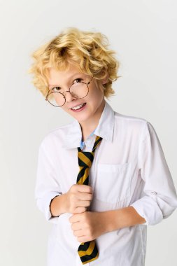 Okul üniformalı sarışın çocuk stüdyo ortamında kravatını tutarken şakacı bir şekilde gülümsüyor..