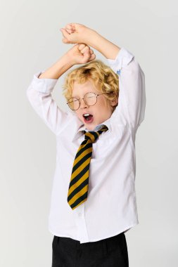 Sarışın bir çocuk stüdyo ortamında okul üniformasını giyerken esniyor..