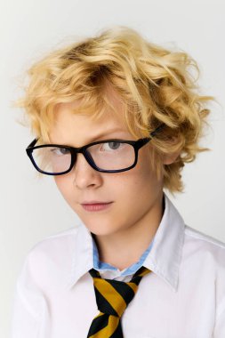 Dalgalı sarı saçlı genç bir çocuk gözlük takıyor ve çizgili kravat takıyor. Stüdyoya güven saçıyor..