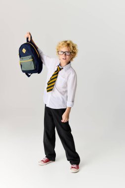 Okul üniformalı neşeli sarışın bir çocuk sırt çantası tutarken gülümsüyor..