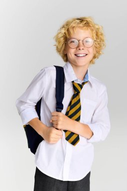 Dalgalı saçlı neşeli sarışın bir çocuk okul üniforması giyiyor ve gülümserken bir çanta tutuyor..