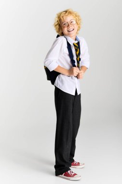 Dalgalı sarı saçlı ve çizgili kravatlı çocuk okul üniformasıyla sınıf için mutlu poz veriyor..