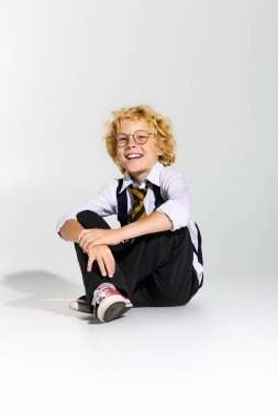 Dalgalı sarı saçlı genç bir çocuk yerde oturuyor, okul üniforması giyiyor ve gülümsüyor..