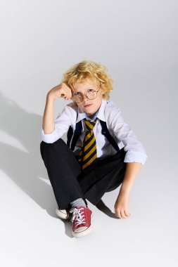 Dalgalı sarı saçlı genç bir çocuk düşünceli bir şekilde okul üniforması içinde oturuyor, çizgili bir kravat takıyor..