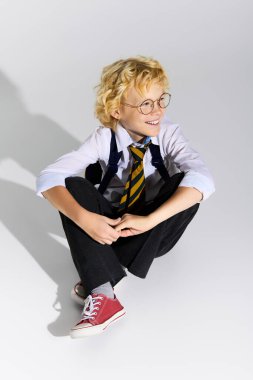 Okul üniformalı neşeli sarışın bir çocuk yerde oturuyor, çizgili kravat ve gözlük takıyor..