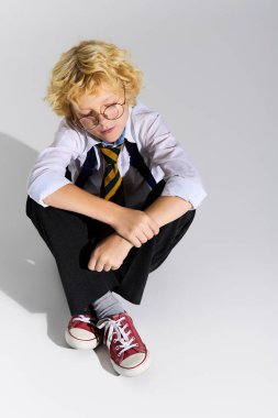 Dalgalı sarı saçlı genç bir çocuk çizgili kravat ve gözlük takmış yerde oturuyor..