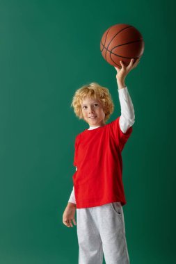 Sarı dalgalı saçlı neşeli bir çocuk elinde basketbol topuyla stüdyoda duruyor..