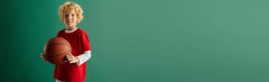 Kırmızı gündelik elbiseli neşeli sarışın bir çocuk elinde basketbol topu ve pankartıyla stüdyoda duruyor.