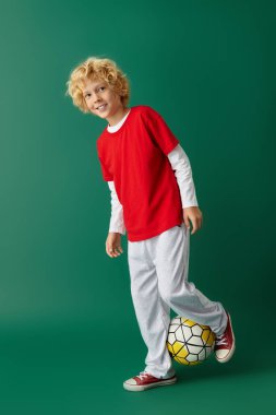 Dalgalı sarı saçlı mutlu bir çocuk futbol topuyla kırmızı bir kıyafet içinde poz veriyor..