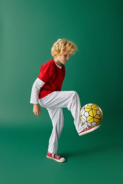 Kırmızılı neşeli sarışın bir çocuk yeşil bir zeminde futbol topunu dengeliyor..