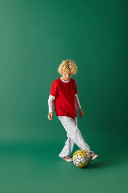 Dalgalı sarı saçlı genç bir çocuk stüdyo ortamında futbol topuyla oynamaktan hoşlanıyor..