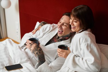 Genç bir çift otel yatağında içkilerini yudumlarken kahkahalar ve sıcaklığı paylaşıyor..