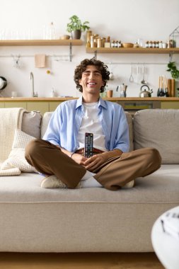 Kıvırcık saçlı yakışıklı genç bir adam koltuğunda rahat bir şekilde oturuyor, uzaktan kumandalı, gülümsüyor..