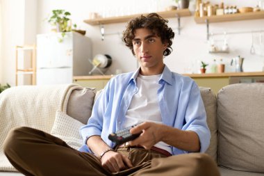 Kıvırcık saçlı yakışıklı genç adam elinde uzaktan kumandayla kanepede rahatlıyor..