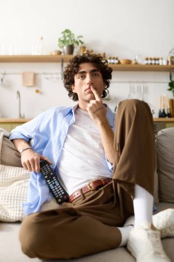 Kıvırcık saçlı genç adam, elinde uzaktan kumandayla derin düşüncelere dalmış, koltuğunda dinleniyor..