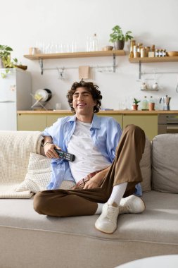 Kıvırcık saçlı genç bir adam evde uzaktan kumandayla dinlenirken gülümsüyor..