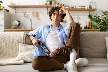 Kıvırcık saçlı genç adam elinde uzaktan kumandayla kanepede rahatlıyor ve evde vakit geçiriyor..