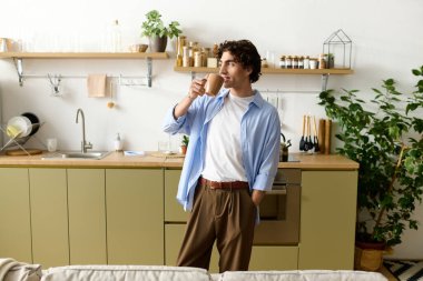 Kıvırcık saçlı genç bir adam şık bir mutfakta duruyor, boş zamanlarında gülümsüyor..