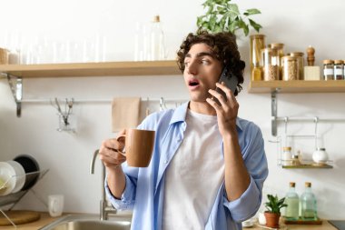 Kıvırcık saçlı genç bir adam evde telefonla konuşurken kahvenin tadını çıkarıyor..