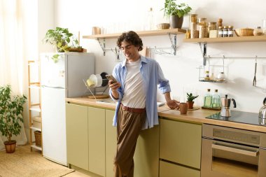 Kıvırcık saçlı genç adam evde rahat anlar geçirir, mutfakta telefonunu kontrol eder..