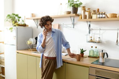 Kıvırcık saçlı genç adam fincandan yudumlar ve mutfakta telefonda sohbet eder..