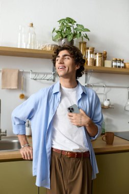 Saçı kıvırcık, yakışıklı genç bir adam ev mutfağında telefonunu tutarken gülümsüyor..