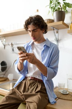 Kıvırcık saçlı genç bir adam evinde rahat bir şekilde oturup telefonuna odaklanıyor..