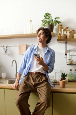 Orta doğulu genç bir adam modern mutfağında rahat bir şekilde oturuyor, gülümsüyor ve telefonunu kullanıyor..