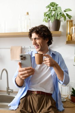 Yakışıklı genç adam elinde bardak tutarken gülümsüyor ve rahat dairesinde telefonuna bakıyor..