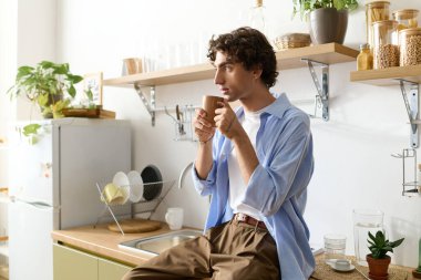 Kıvırcık saçlı genç adam evde dinleniyor, parlak bir mutfakta oturmuş kahvesini yudumluyor..