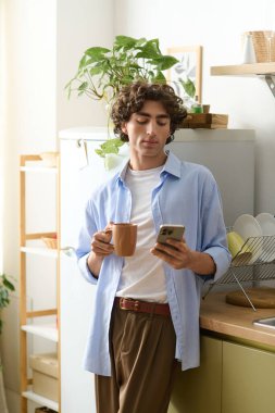 Kıvırcık saçlı yakışıklı genç adam evde rahatlıyor, kahvesini yudumluyor ve telefonunu karıştırıyor..