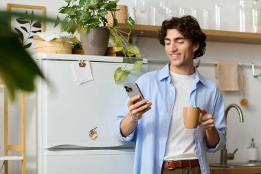 Genç adam elinde kahve fincanı tutarken gülümsüyor ve akıllı telefonuna içeride bakıyor..