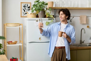 Orta Doğulu genç bir adam, kıvırcık saçlı, elinde kupa ve telefonu olan rahat evinde gülümsüyor..