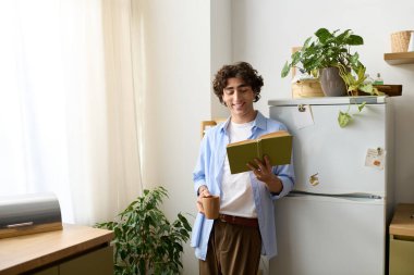 Kıvırcık saçlı genç bir ortadoğulu evde sıcak bir şeyler içerken kitap okuyor..