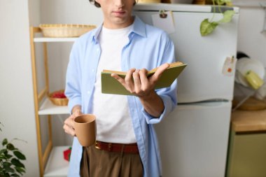 Genç adam rahat evinde kitap ve sıcak bir içecekle rahatlıyor..