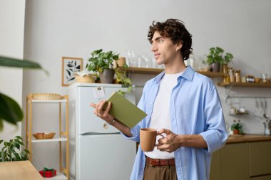 Genç, kıvırcık saçlı, orta doğulu bir adam dairesinde kitap ve sıcak bir içecek tutuyor..
