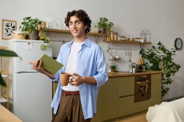 Kıvırcık saçlı yakışıklı genç bir adam dairesinde rahatlıyor, kitap okurken bir şeyler içiyor..