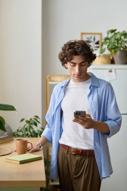 Genç adam evde dinleniyor, elinde sıcak bir içecek tutarken telefonuyla kayıyor, etrafı bitkilerle çevrili..