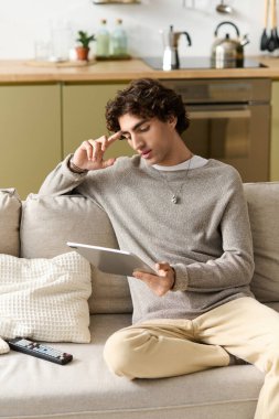 Genç adam evdeki kanepede dinlenirken tablet kullanıyor ve derin derin düşünüyor..