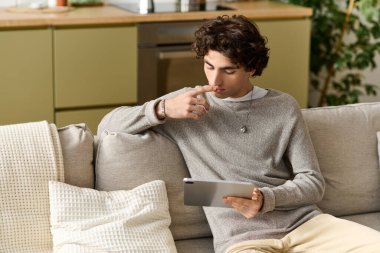 Kıvırcık saçlı yakışıklı bir genç adam evde eğleniyor, tabletine odaklanıyor..