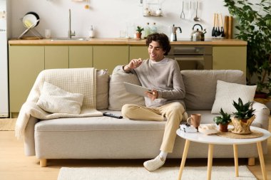 Kıvırcık saçlı yakışıklı genç bir adam, şık bir dairede tablet kullanarak kanepede dinleniyor..