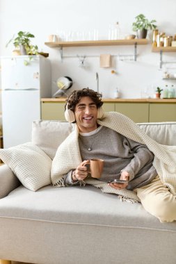 Orta Doğulu kıvırcık saçlı bir adam sıcak içecek ve müziğin tadını çıkarırken koltukta dinleniyor..