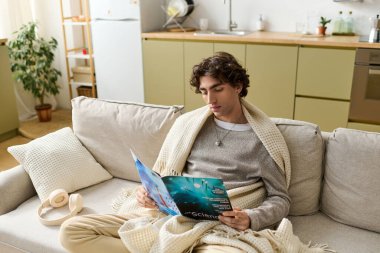 Kıvırcık saçlı genç bir adam, evinde dergi okumaya dalmış, koltukta dinleniyor..