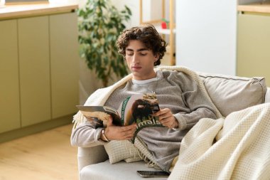 Kıvırcık saçlı genç bir adam evde vakit geçirmekten hoşlanıyor, kanepede dergi okuyor..