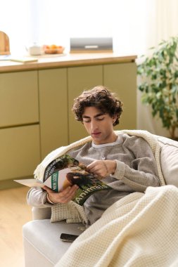 Kıvırcık saçlı adam, sıcak, davetkar bir mekanla çevrili bir dergi okurken kanepede dinlenir..